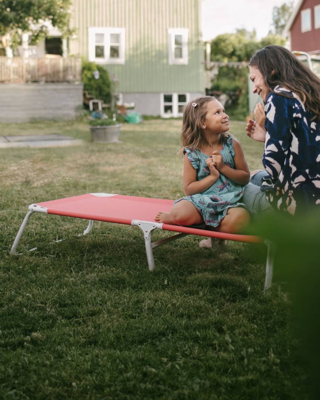Ett barn och en kvinna sitter och pratar på en solstol i en somrig trädgård.